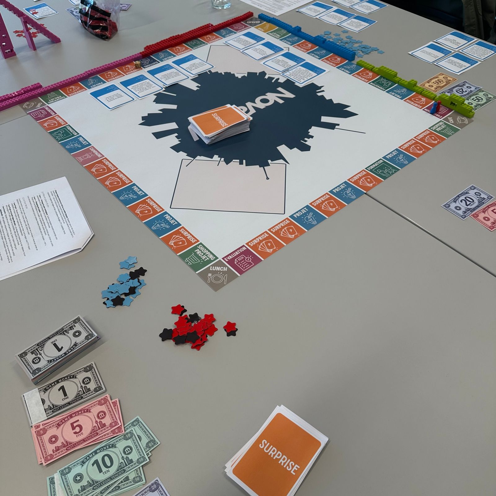 Vue du plateau avec la pile de cartes Surprise au centre, des cartes Projet et des billets de jeu.